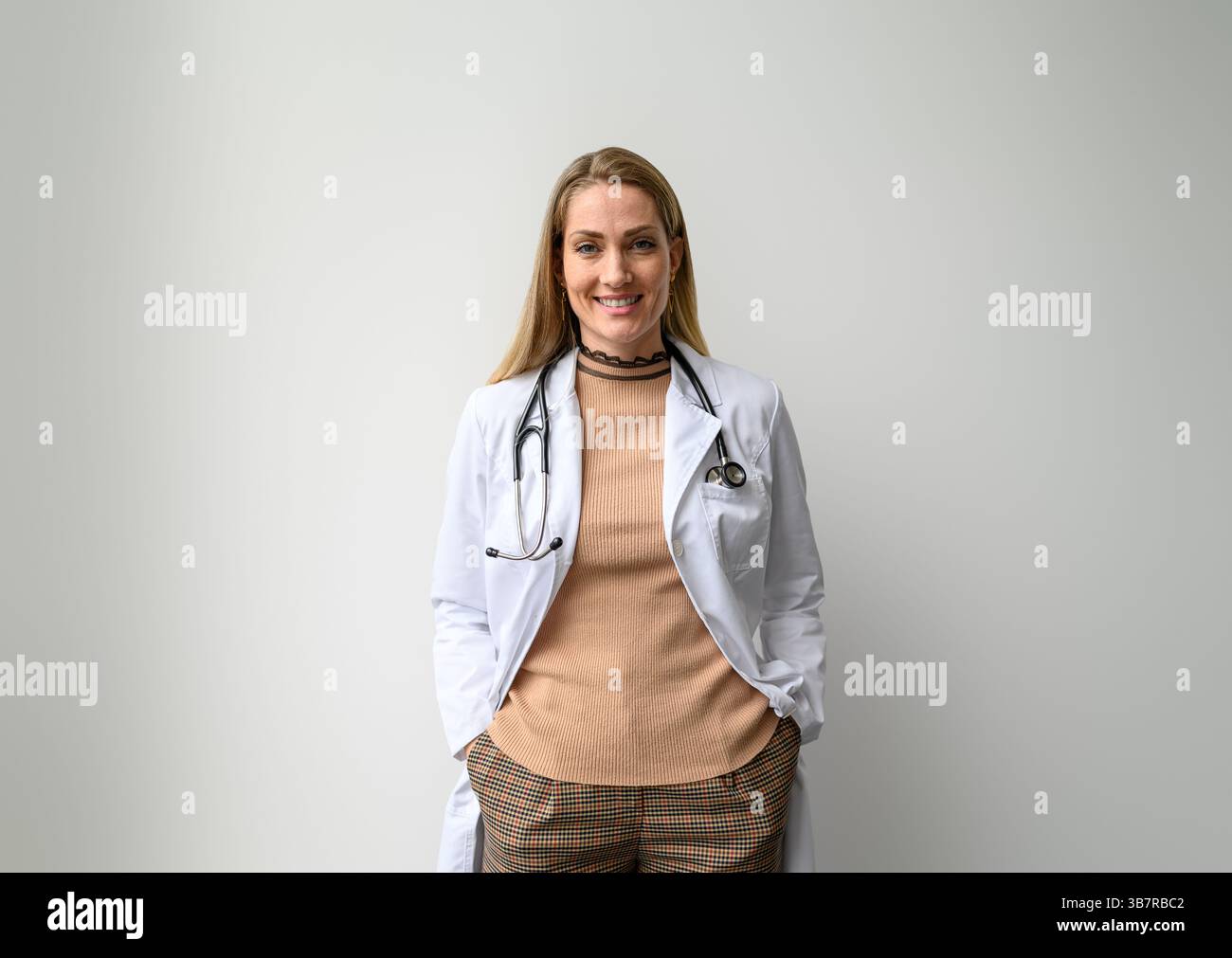 Ritratto di donna medico professionista in camice da laboratorio e con le mani in tasche in piedi su sfondo bianco Foto Stock