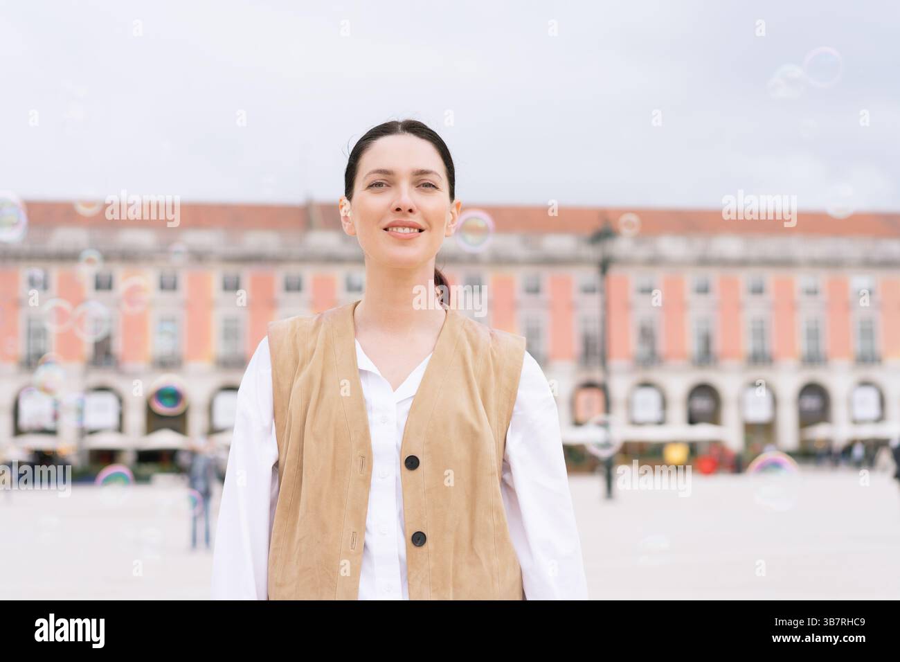 Donna sorridente in gilet marrone in piedi in piazza mentre guarda la macchina fotografica. Una donna turistica che posa di fronte a un edificio sfocato, trasuda calore e fiducia in un ambiente urbano Foto Stock