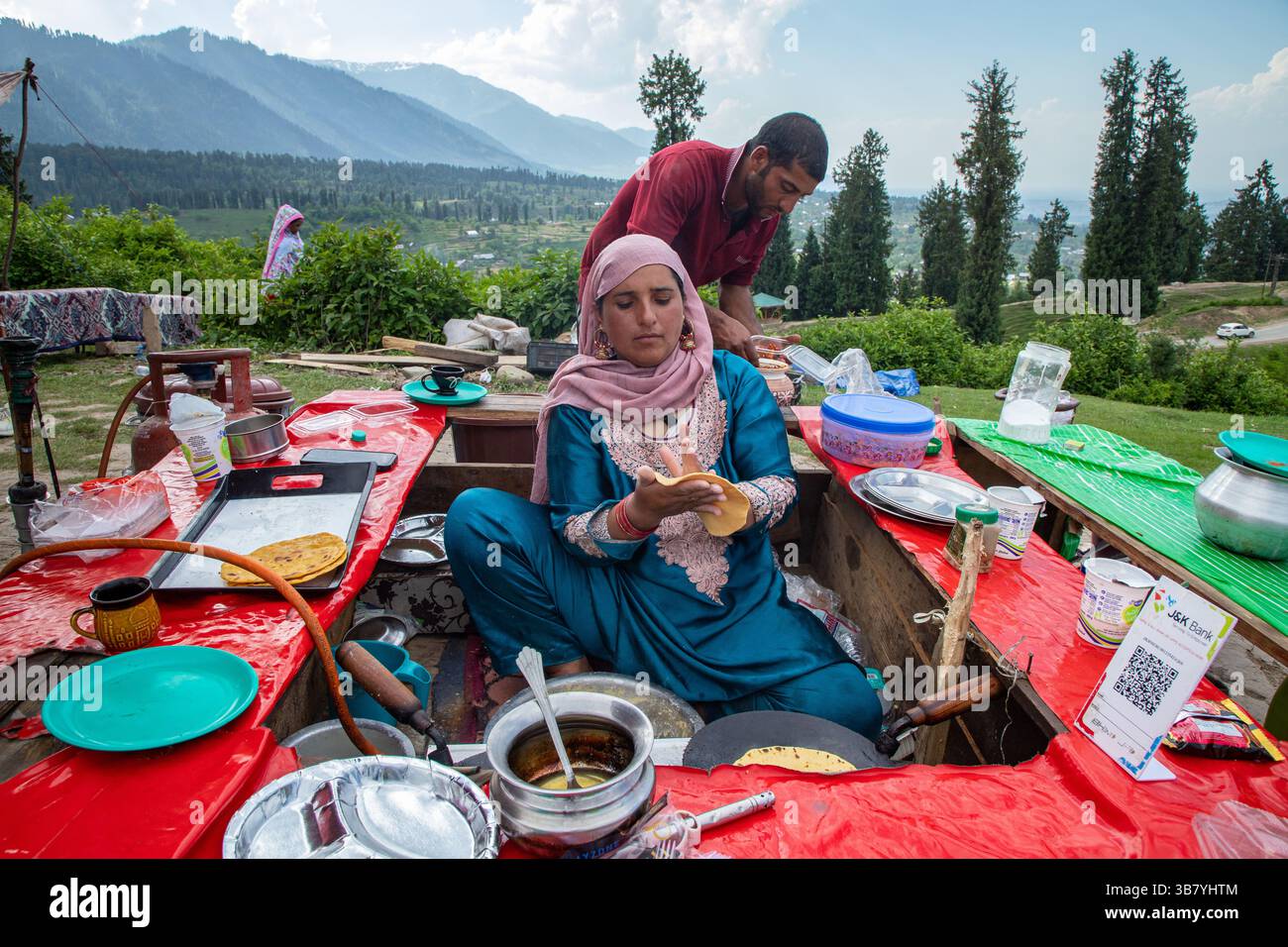 23 maggio 2024, Doodhpathri, Jammu e Kashmir, India: Una donna del Kashmir prepara tortillas di farina di mais mentre vende Noon Chai (tè salato) ai turisti e alla gente del posto passando per la sua banchina lungo la strada Doodhpathri nel distretto di Budgam, a sud-ovest di Srinagar. Le autorità hanno chiesto ai proprietari dei venditori di tè di liberare lo spazio che hanno occupato ''illegalmente'' entro dieci giorni o di affrontare azioni. I funzionari hanno detto a un'agenzia di stampa locale che l'autorità per lo sviluppo del turismo ha ricevuto numerose lamentele per il fatto che i proprietari delle bancarelle di tè hanno tagliato rami di alberi, danneggiando così le immagini Foto Stock