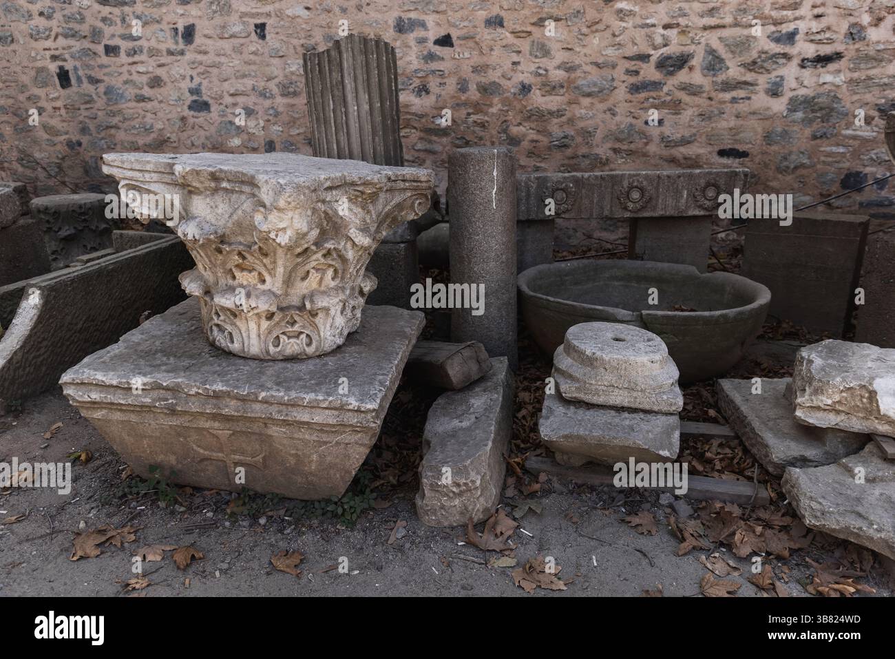Una collezione di antichi manufatti in pietra, tra cui colonne e capitelli, disposti contro un muro di mattoni intemprati in un ambiente esterno, circondato da Foto Stock Una collezione di antichi manufatti in pietra, tra cui colonne e capitelli, disposti contro un muro di mattoni intemprati in un ambiente esterno, circondato da Foto Stock