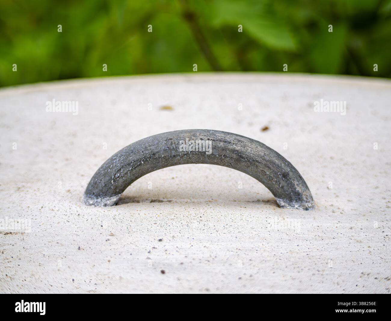 Primo piano di un manico metallico su una superficie in cemento, circondato dal verde. L'impugnatura è leggermente curva e mostra segni di usura, in contrasto con Foto Stock
