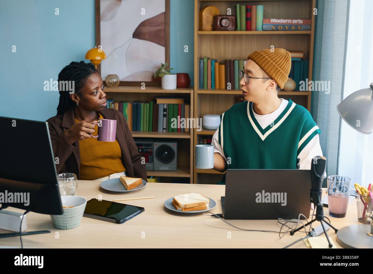 Donna afroamericana e uomo asiatico seduti ad un tavolo di legno con tazze da caffè e piatti da colazione, impegnati in una discussione in un accogliente ufficio domestico Foto Stock