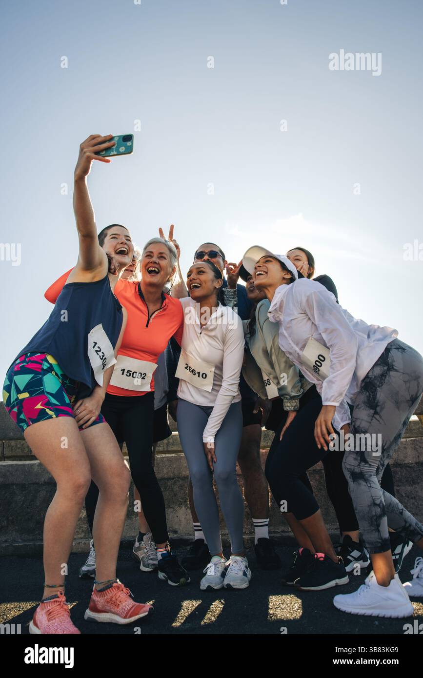 Un gruppo di amici di un club di running si riunisce per un allegro selfie durante un evento all'aperto. Indossati con abbigliamento atletico e pettorali da corsa, irradiano happ Foto Stock