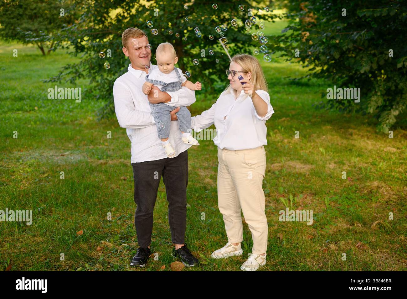 Il fine settimana, la famiglia si rilassa nel parco. Mamma che soffia bolle di sapone, papà che tiene il bambino in braccio, si diverte, ridendo. Famiglia che cattura le bolle, gioca insieme Foto Stock