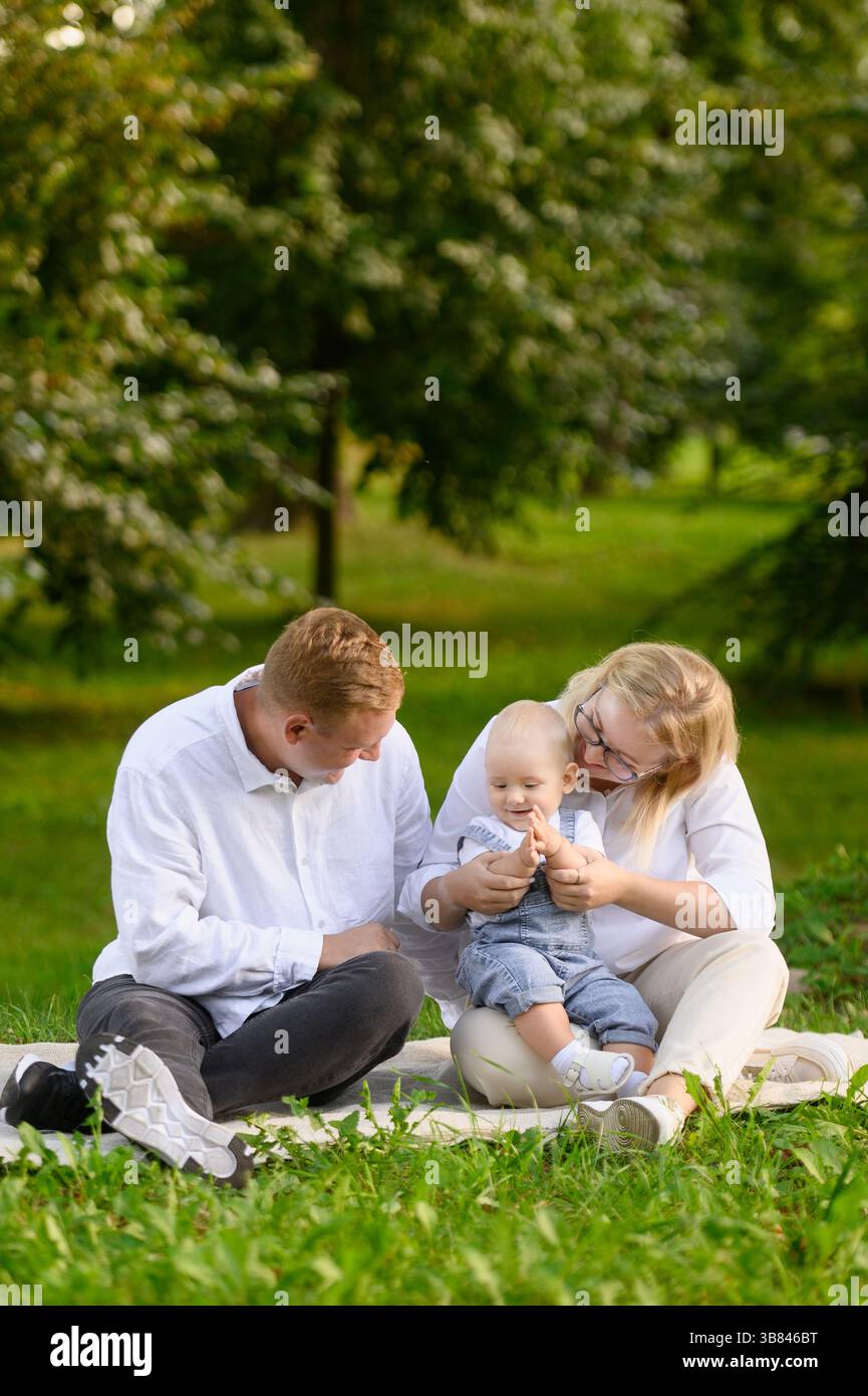 Felice coppia con bambino seduto su una coperta nel parco durante le vacanze, mamma che tiene il bambino in braccio e gioca con lui una torta di pappy, papà felicemente guardando t Foto Stock