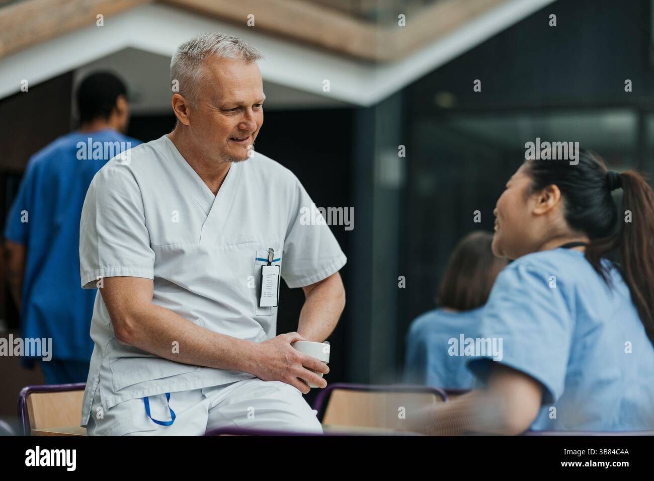 Medico maschio maturo che parla con il collega mentre si siede sul tavolo in mensa dell'ospedale Foto Stock