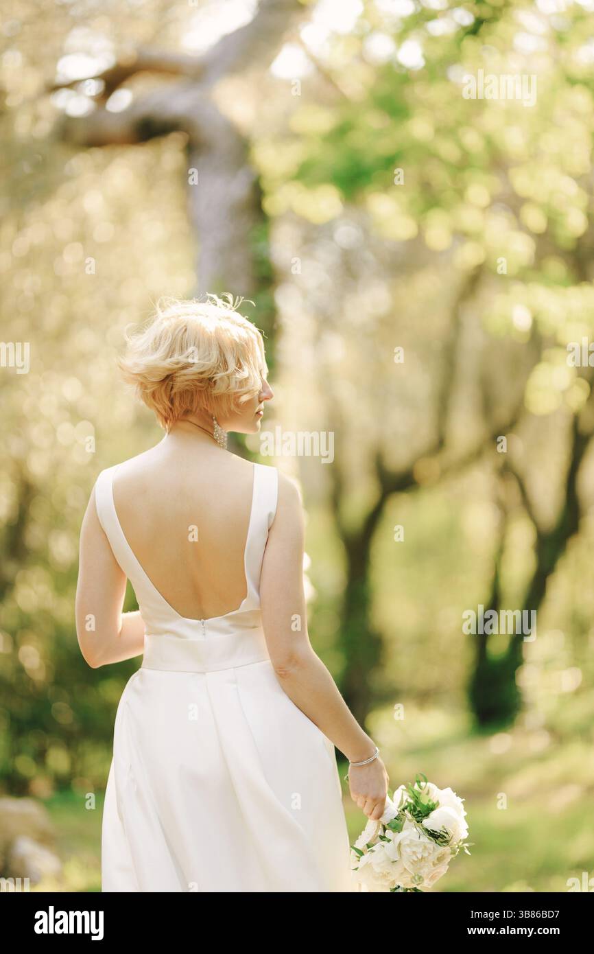 La sposa in un abito bianco con un mazzo di fiori tra le mani si trova nel parco con la testa girata a destra. Foto Stock