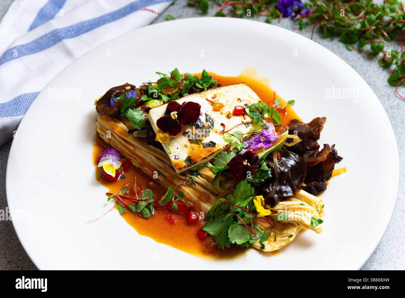 Tofu splendidamente placcato con funghi auricolari in legno, enoki, fiori commestibili ed erbe aromatiche serviti in una ricca salsa d'arancia su piatto bianco. Foto Stock