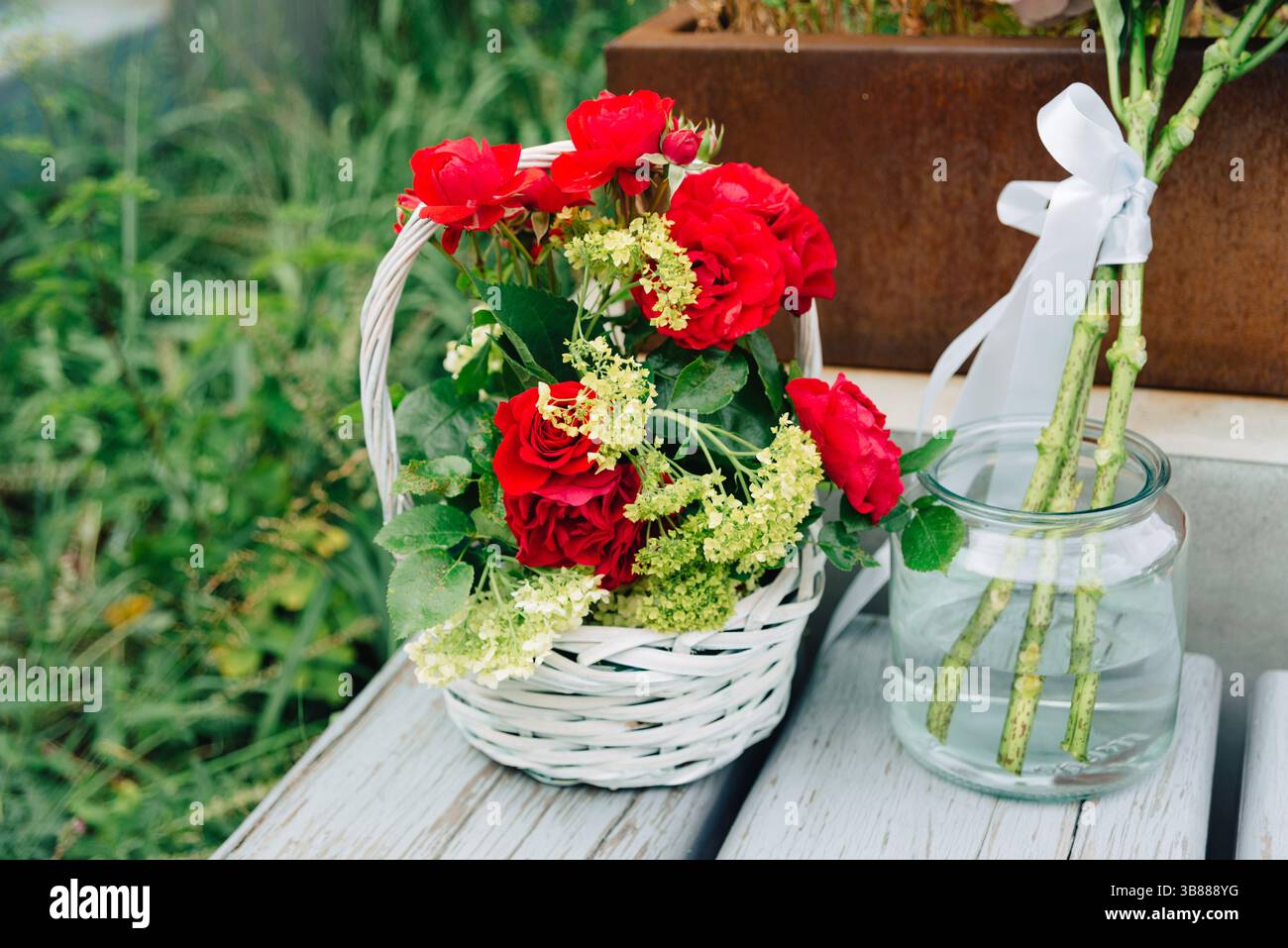 Un cestino di vimini bianco pieno di rose rosse e vegetazione si trova su una panchina resistente agli agenti atmosferici accanto a un vaso con gambi di rose e nastro. Foto Stock