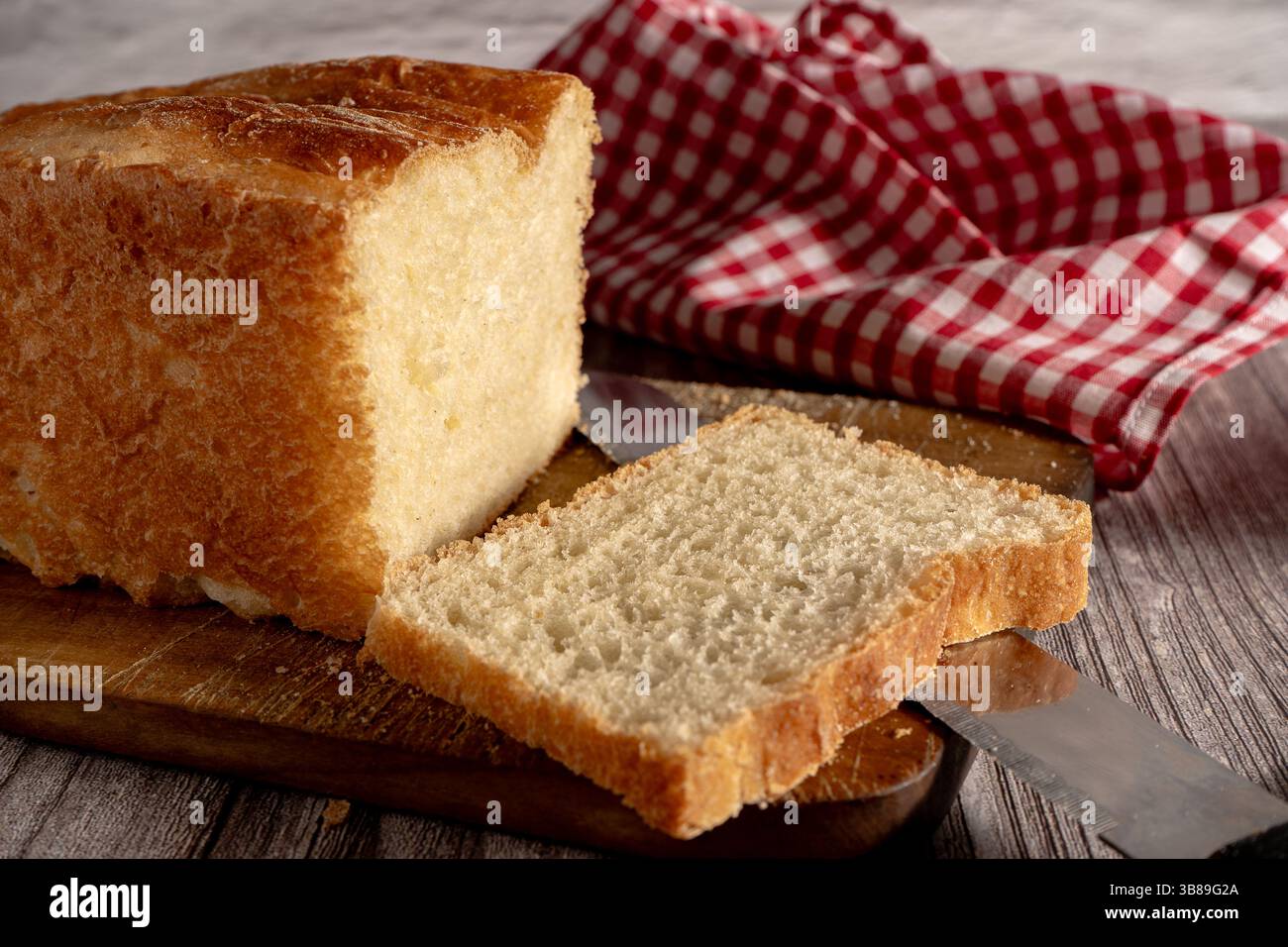 Pane appena sfornato con una fetta tagliata su una tavola di legno e un panno a scacchi rosso Foto Stock