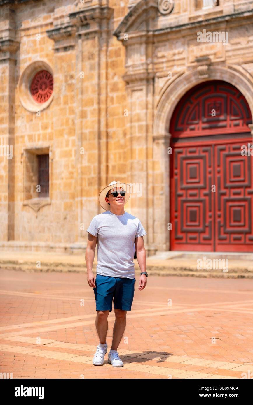 Sorridente giovane latino in abiti leggeri e casual e cappello di paglia in piedi di fronte alla storica chiesa in pietra con porta rossa a Cartagena, Colombia. Foto Stock