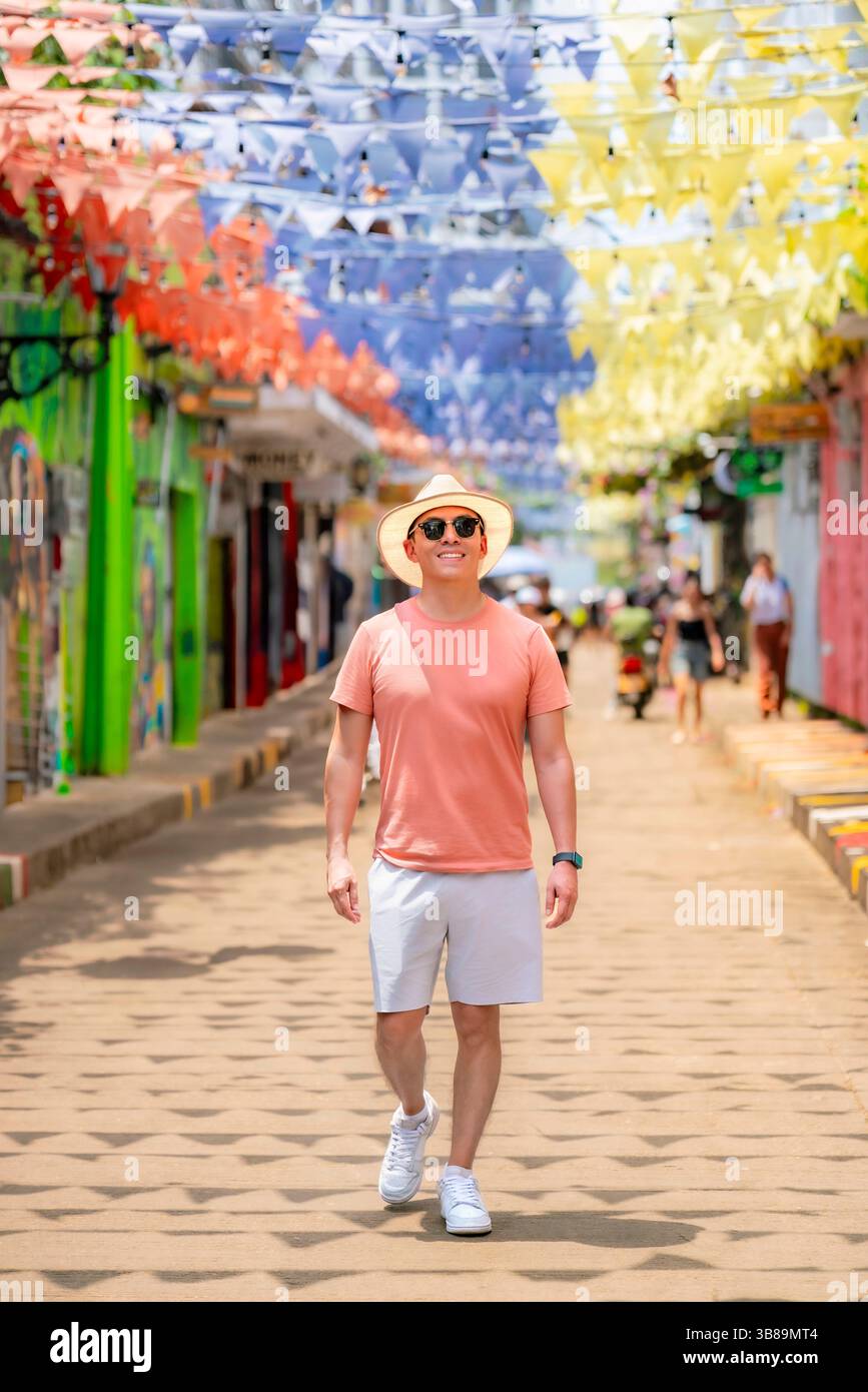 Il giovane latino sorridente cammina lungo una strada decorata a Getsemaní, Cartagena, indossando camicia in corallo, pantaloncini bianchi, cappello di paglia e occhiali da sole Foto Stock