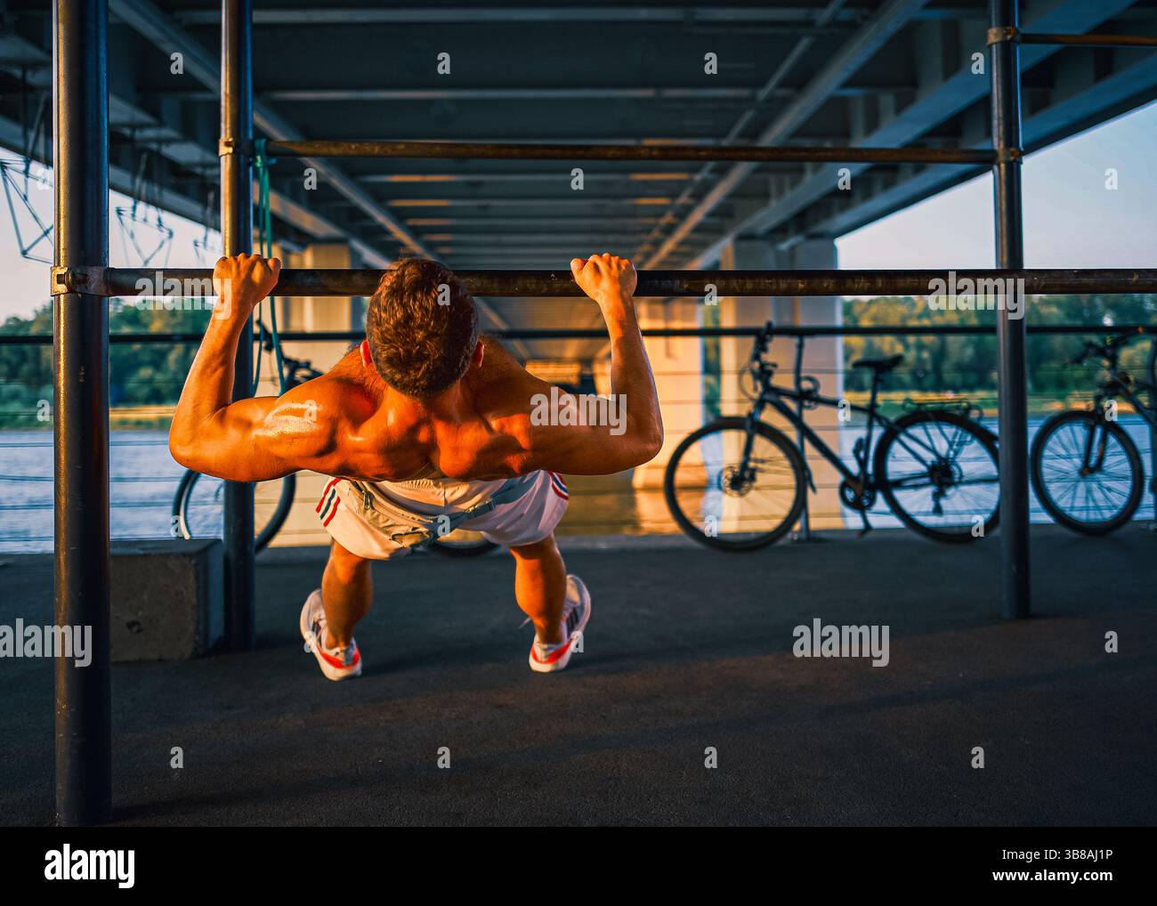 Un uomo muscoloso si allena sotto un ponte utilizzando barre orizzontali per esercizi di peso corporeo. L'intensa luce dorata del tramonto mette in risalto la sua forma atletica. Foto Stock