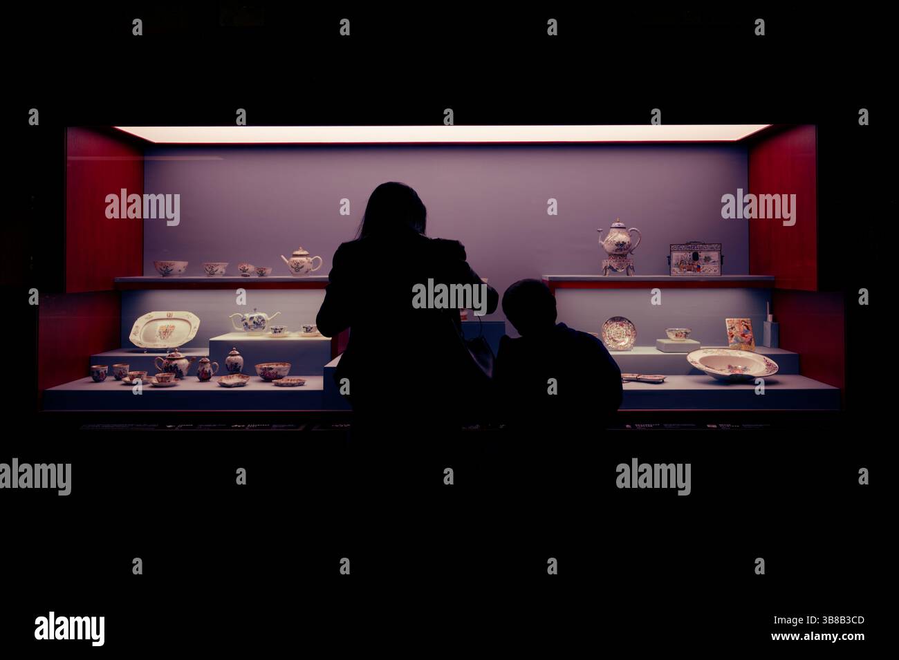 Le sagome di due visitatori ammirano un'esposizione illuminata di teiere e stoviglie in porcellana ornate Foto Stock