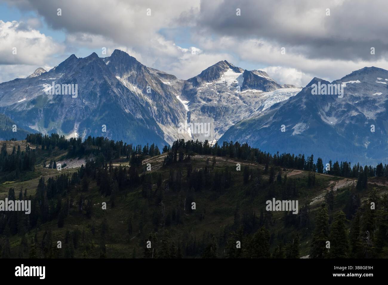 Alte montagne si innalzano maestosamente sullo sfondo di nuvole sparse, mentre un vivace paesaggio verde riempie il primo piano. La scena mostra nat Foto Stock