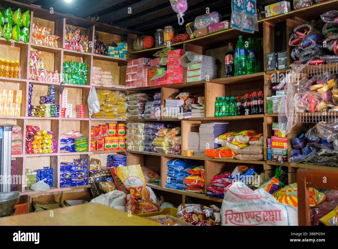 Un negoziante rifornisce gli scaffali con spuntini, bevande e articoli per la casa per i clienti nel loro piccolo negozio generale. Foto Stock