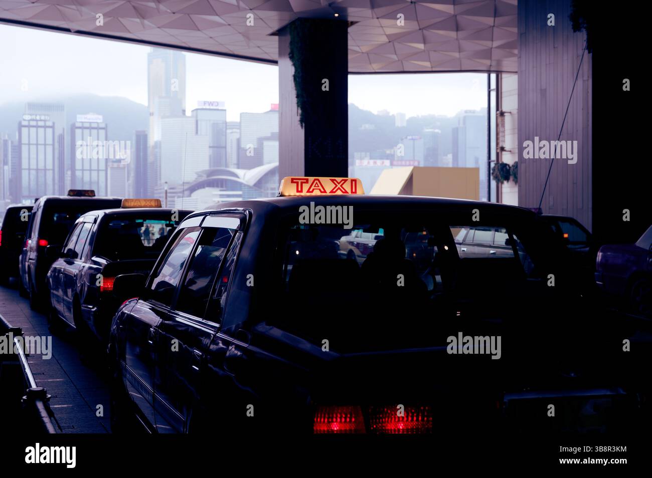 La coda dei taxi sotto la tettoia geometrica con lo skyline di Hong Kong sullo sfondo Foto Stock