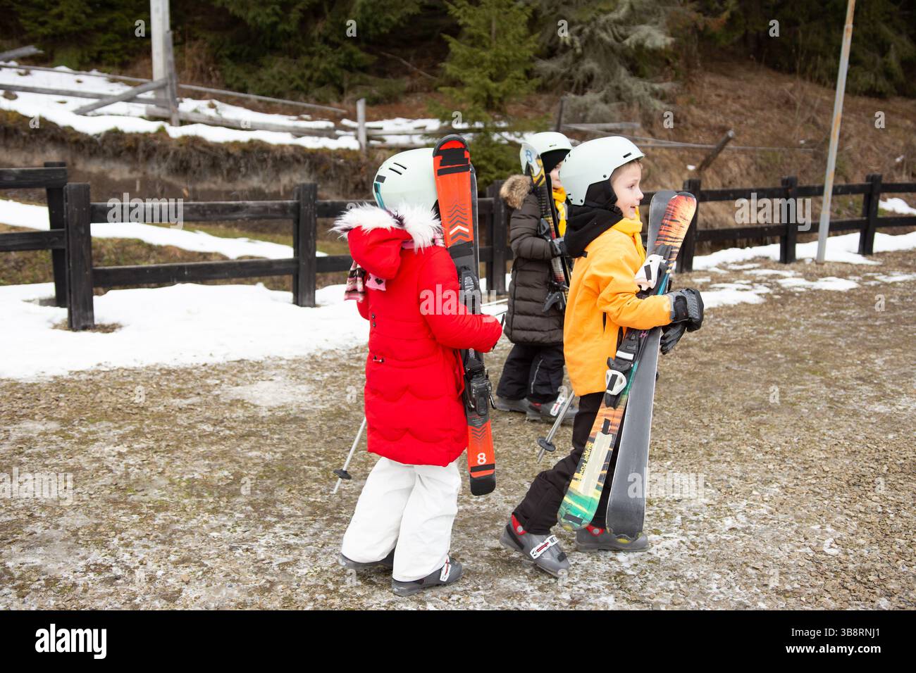 I bambini in abiti da sci vivaci portano gli sci mentre si dirigono verso le piste. La neve splende sullo sfondo, suggerendo una giornata piena di divertimento Foto Stock