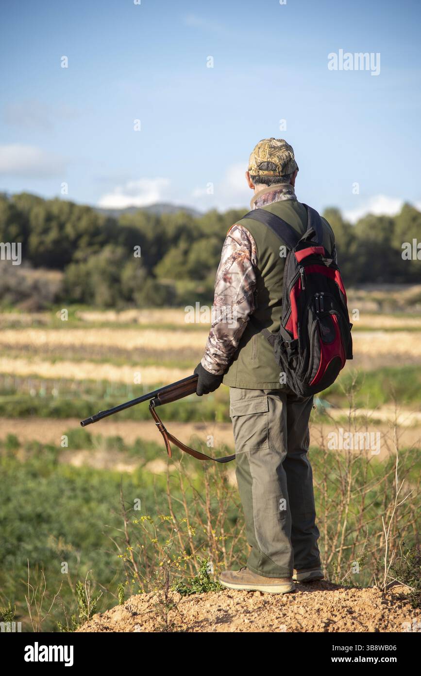 Cacciatore che indossa un mimetismo e tiene il fucile in piedi su una piccola collina che si affaccia su un campo durante una caccia diurna a Sant Pere de Ribes, Barcellona, Spa Foto Stock