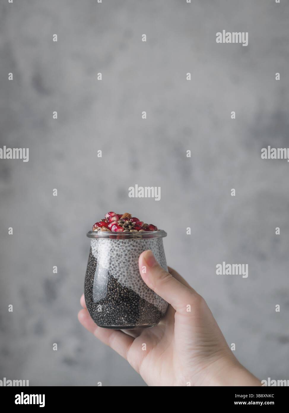 Budino di Chia con carbone nero e latte bianco di vaniglia a base vegetale in mano da donna su sfondo grigio. Vaso in vetro femmina con impugnatura a mano e servomeccanismo chia puding Foto Stock
