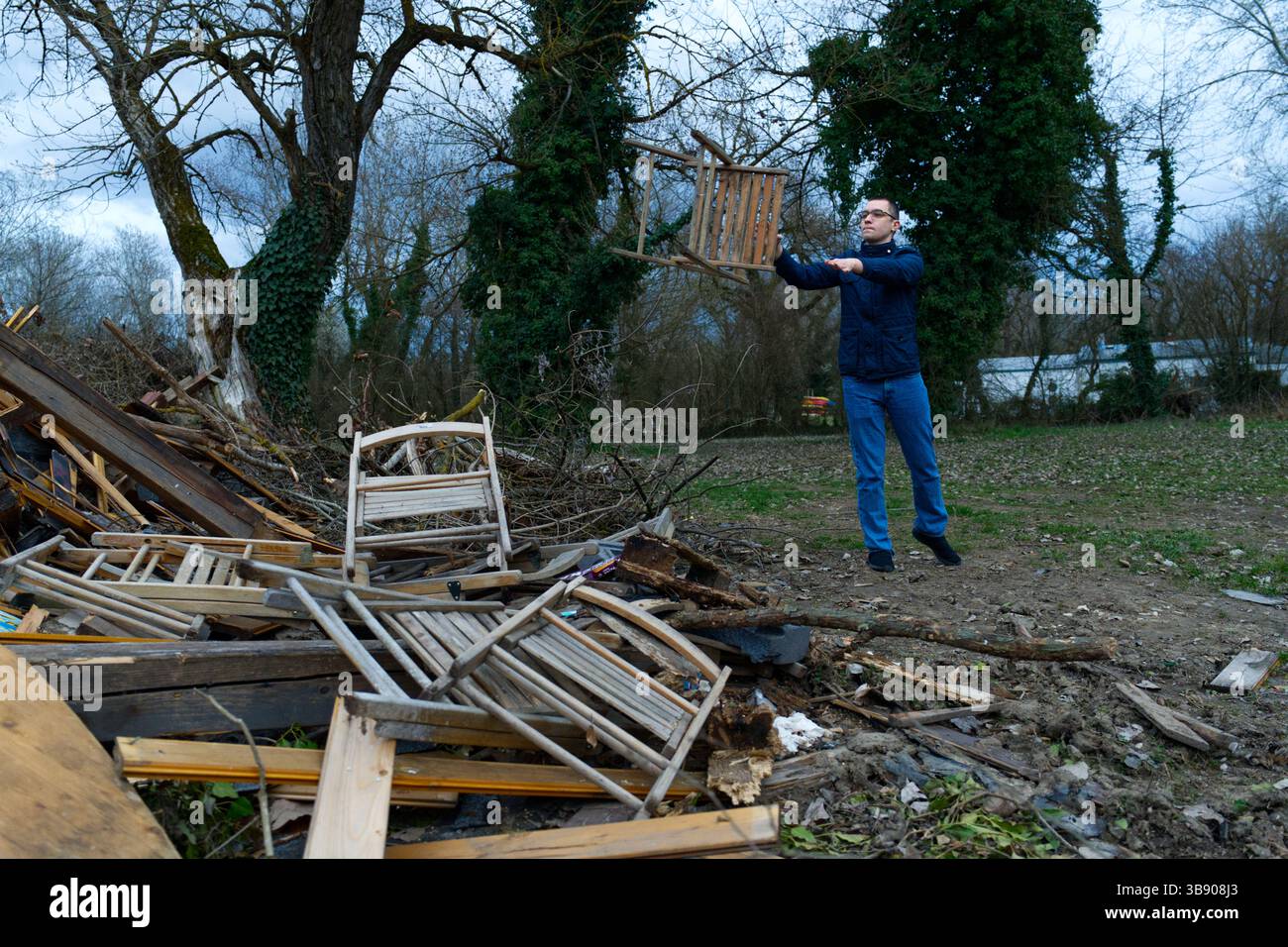 Un uomo lancia una sedia di legno tra un mucchio di mobili rotti in un ambiente esterno desolato. La scena suggerisce l'abbandono o la pulizia dopo un evento. Foto Stock