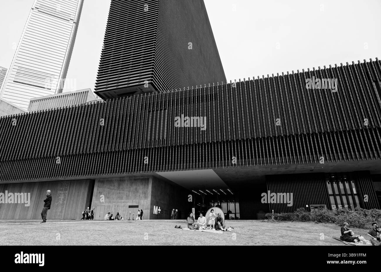 Linee architettoniche sorprendenti della torre M+ Museum sopra un tranquillo momento familiare sul piazzale erboso Foto Stock
