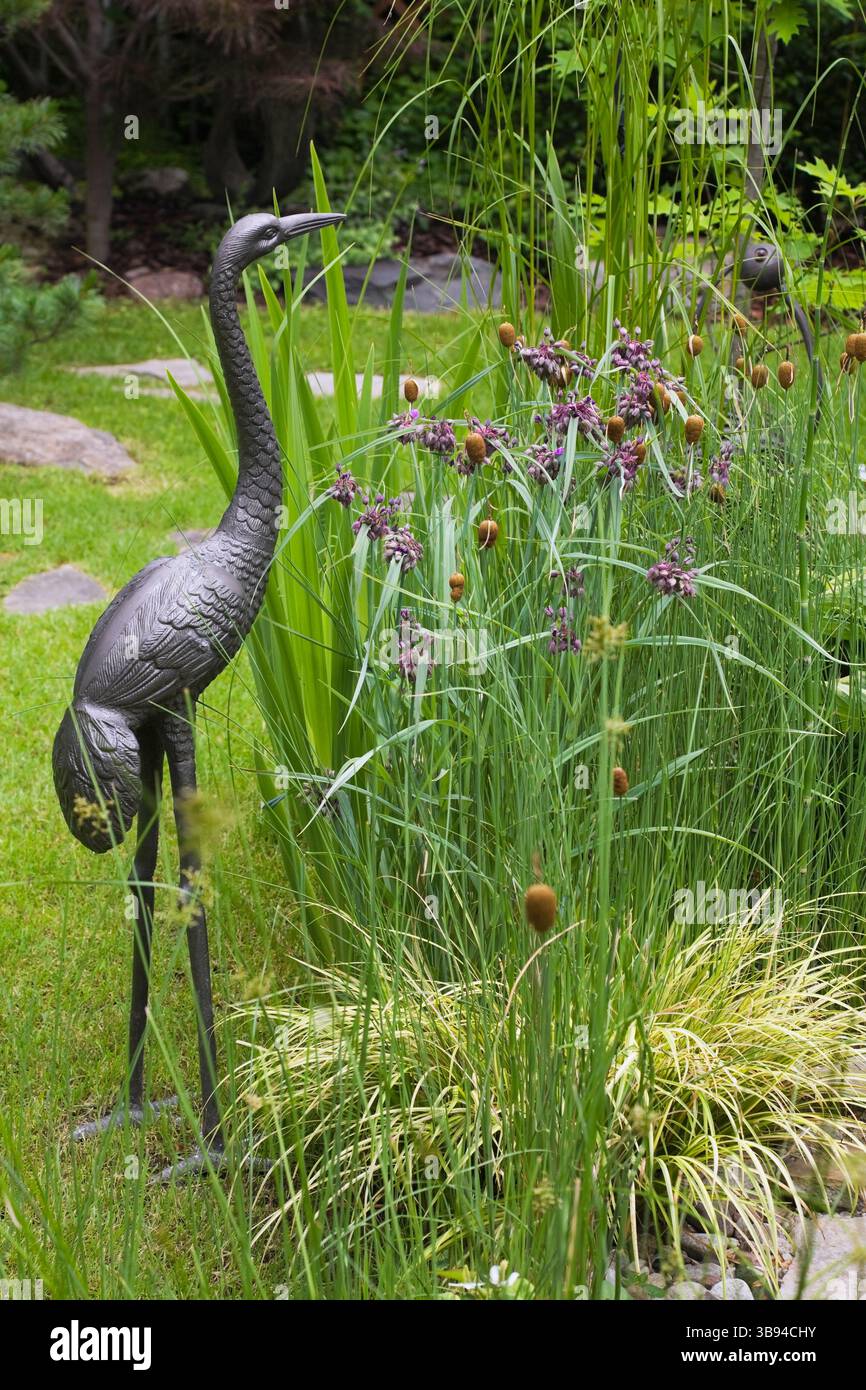 Scultura di uccello d'Heron in metallo e fiori viola di Tradescantia virginiana - verga di Spiderwort in bordo misto nel giardino sul retro in estate. Foto Stock