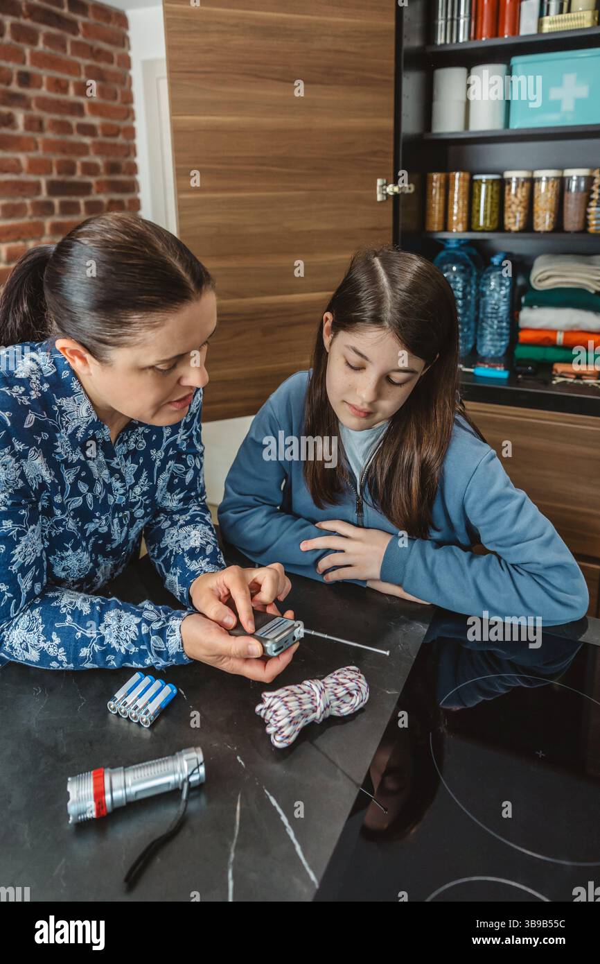 Madre e figlia ascoltano notizie di emergenza in un piccolo ricevitore radio portatile durante il blackout elettrico. Famiglia pronta con dispensa alimentare di emergenza in k Foto Stock