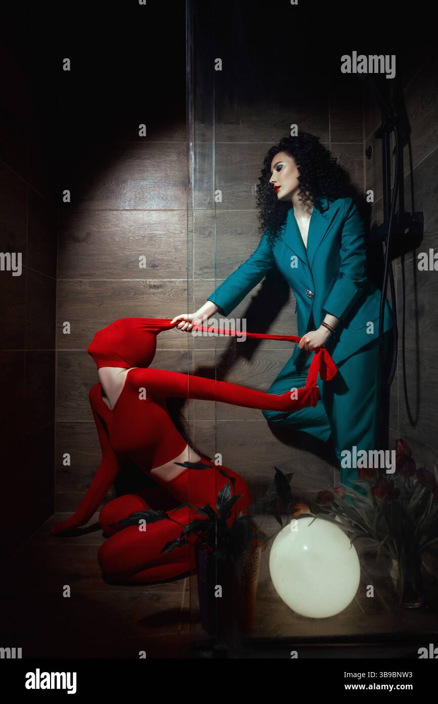 Un'immagine potente di movimento e controllo: Una donna in verde acqua tira una figura in costume rosso in uno studio oscuro. Tesa, metaforica e artistica. Foto Stock