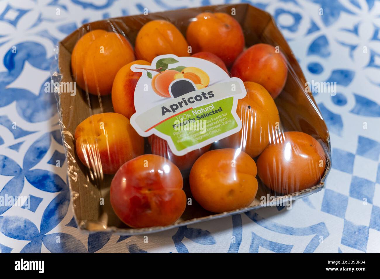 Primo piano di albicocche mature confezionate ordinatamente in un vassoio di cartone avvolto in pellicola di plastica. La frutta colorata e l'etichetta contrastano splendidamente con il blu Foto Stock