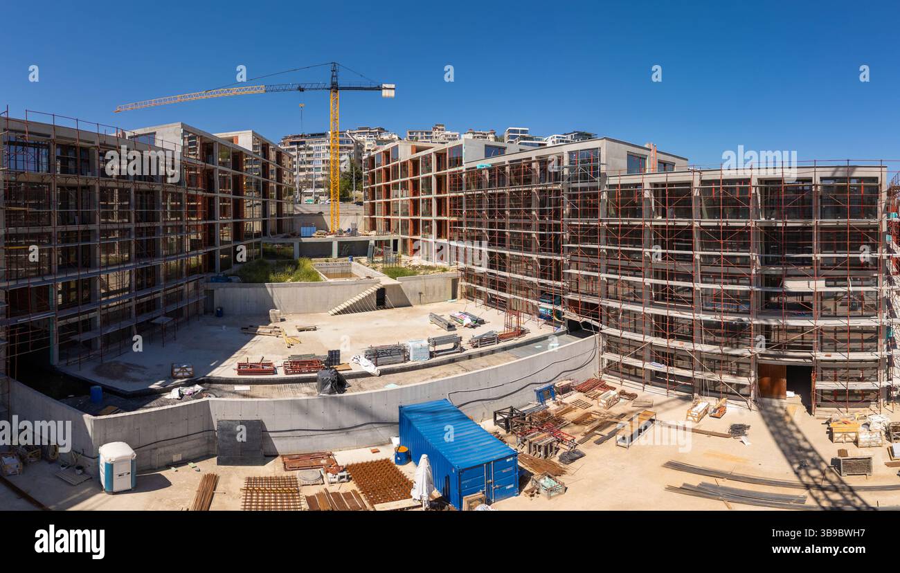 Vista panoramica di un moderno cantiere residenziale con impalcature, gru ed edifici incompiuti sotto un cielo azzurro limpido in una città Foto Stock