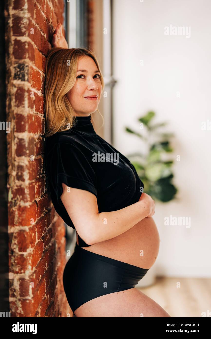 Donna incinta in nero appoggiata contro le pareti di mattoni in casa Foto Stock