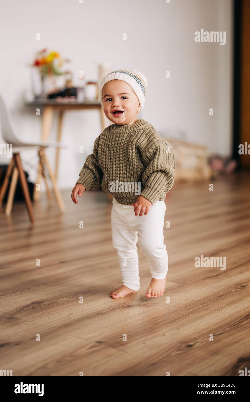 Bimbo sorridente in maglia e berretto che cammina in casa Foto Stock