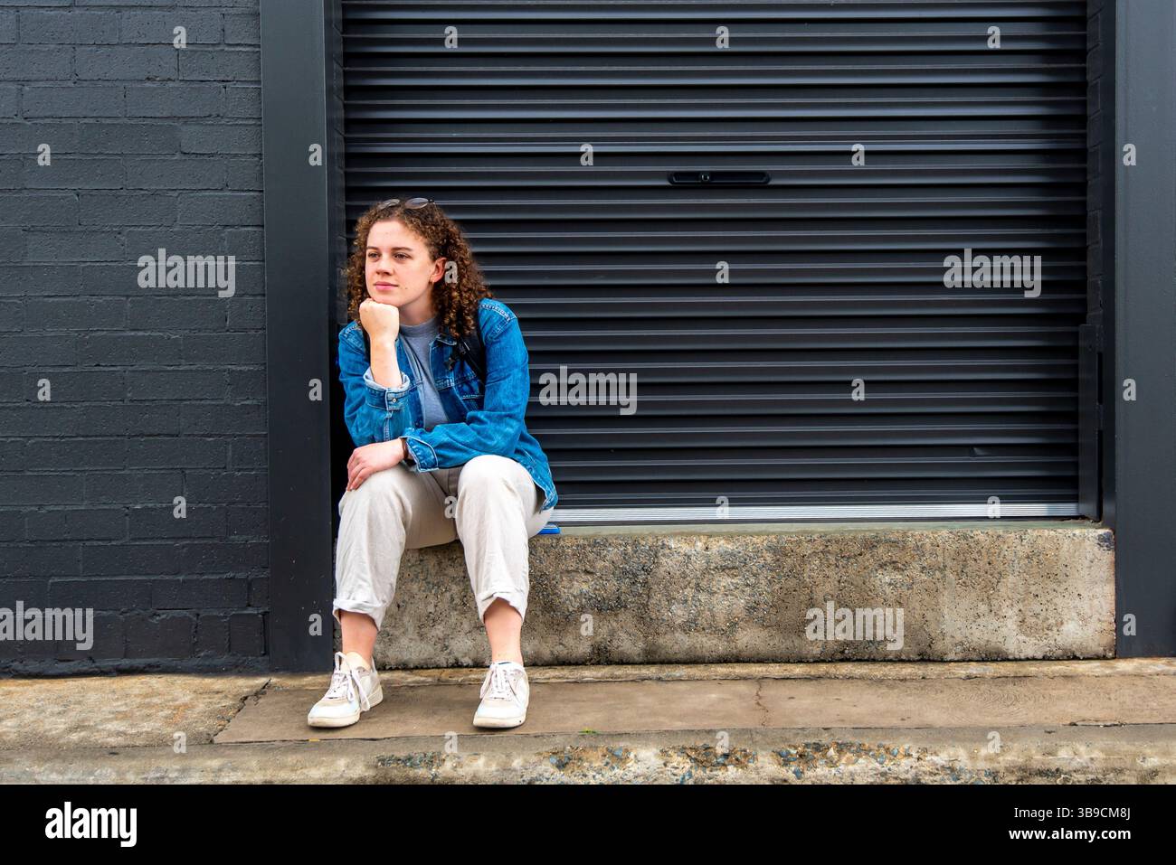 Una giovane donna si siede su un passo concreto che le regge il mento sul braccio con gli occhiali da lettura appoggiati sulla fronte, contemplando la vita Foto Stock