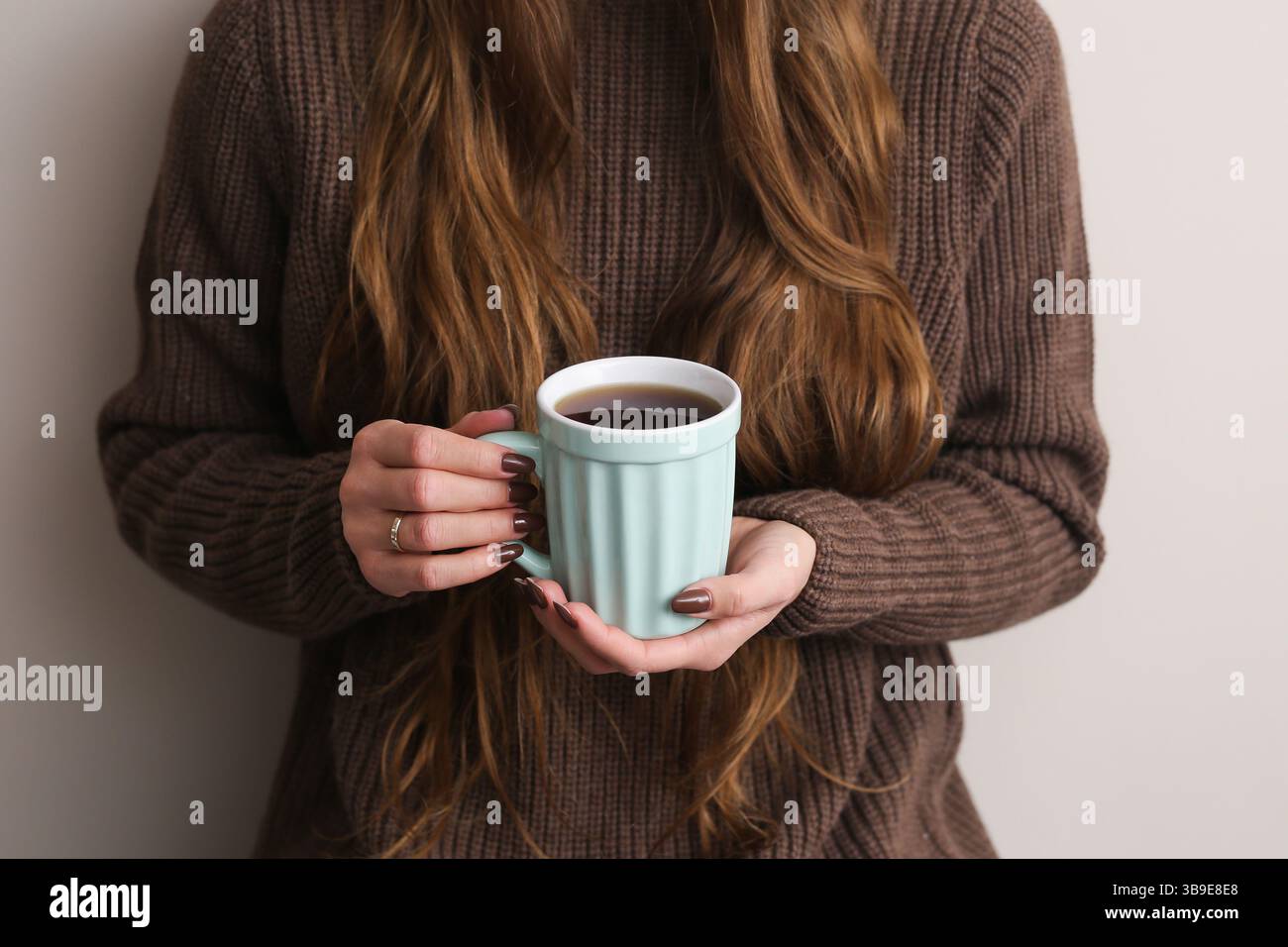 Donna che tiene una tazza pastello. Accogliente mattina autunnale al coperto. Bevanda calda in mano. Maglione marrone e capelli lunghi. Un tranquillo momento di stile di vita hygge. Foto Stock