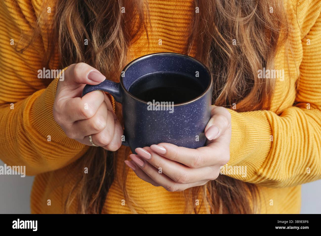 Donna che tiene la tazza da caffè. Bevanda nera calda in mano. Maglione giallo e capelli lunghi. Caffè caldo e accogliente. Mattinata rilassata sullo sfondo dello stile di vita di casa Foto Stock