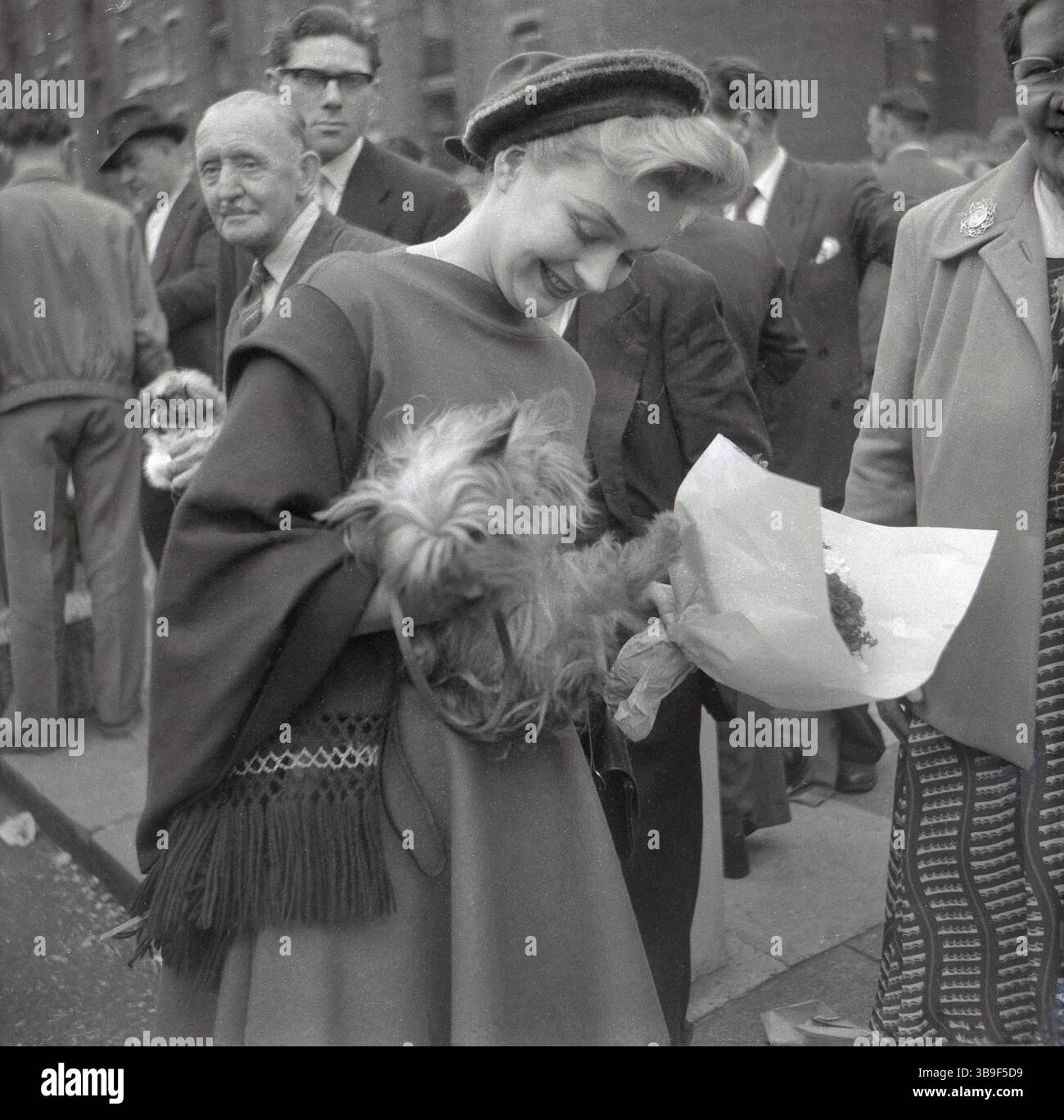 Anni '1950, storica e attraente giovane signora, in abbigliamento elegante al club Row East End Market con un piccolo cane carino, Londra, Inghilterra, Regno Unito. Il mercato all'aperto della fila dei club nell'East End di Londra, così come l'offerta tradizionale di abbigliamento, articoli per la casa, giocattoli e cibo, aveva anche animali in vendita. Foto Stock