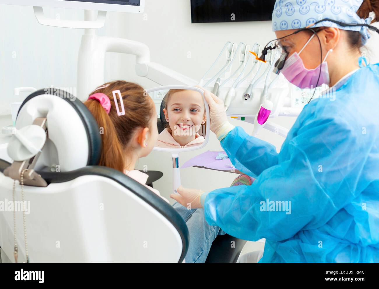 Il dentista pediatrico mostra uno specchio a una ragazza sorridente seduta su una sedia dentista, permettendole di vedere i risultati della procedura dentale in una clinica moderna Foto Stock