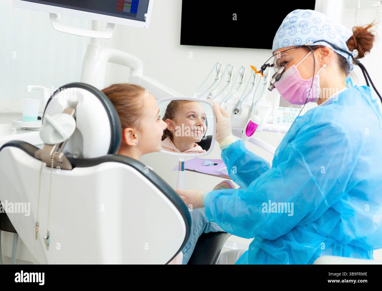 Il dentista pediatrico mostra uno specchio a una ragazza sorridente seduta su una sedia dentista, permettendole di vedere i risultati della procedura dentale in una clinica moderna Foto Stock