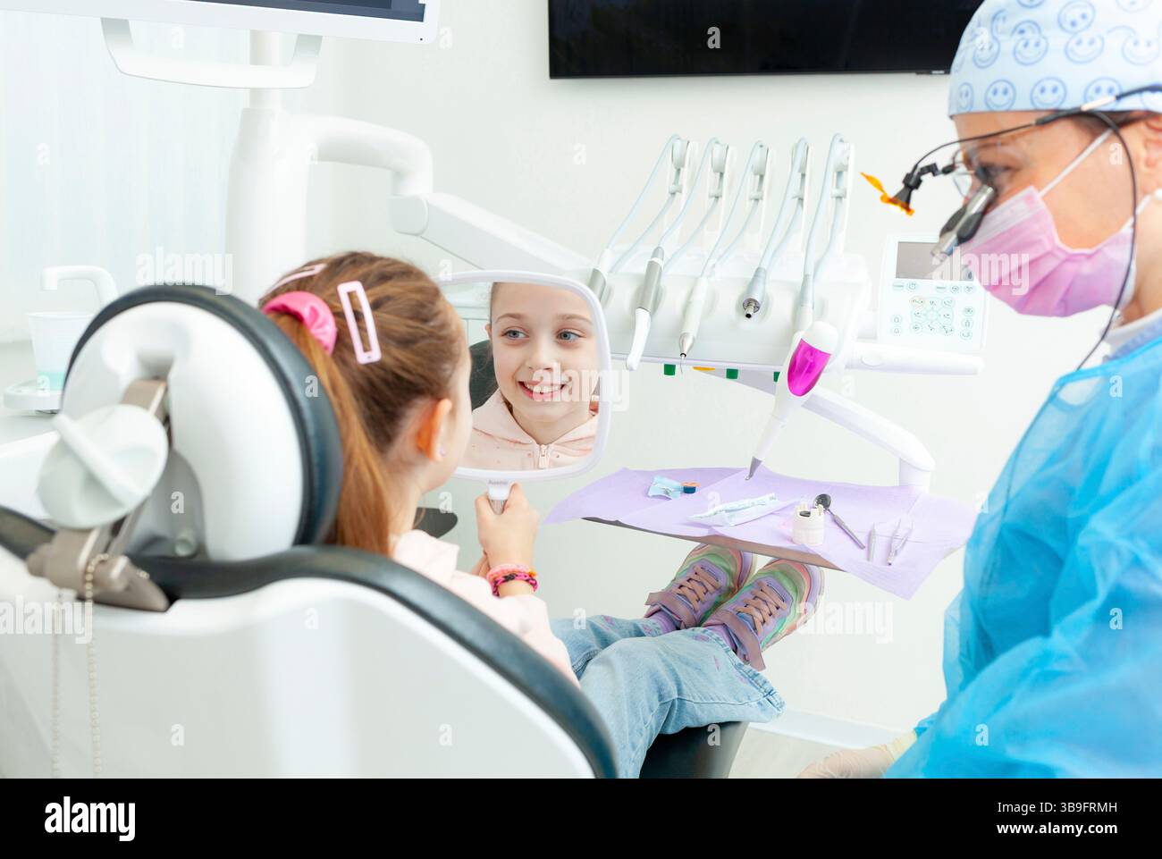 Il dentista pediatrico mostra uno specchio a una ragazza sorridente seduta su una sedia dentista, permettendole di vedere i risultati della procedura dentale in una clinica moderna Foto Stock