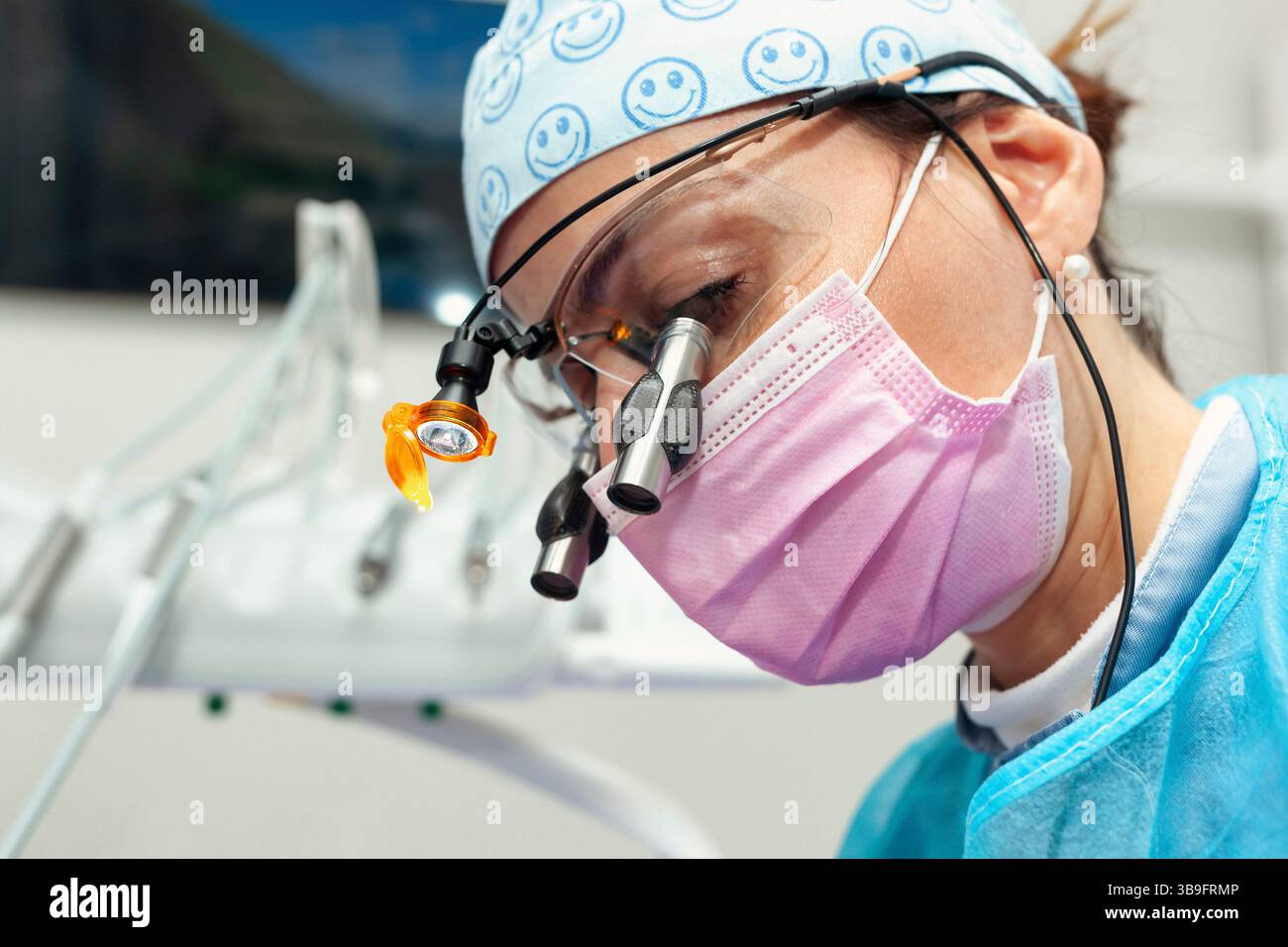Dentista donna che indossa un berretto chirurgico, lenti di ingrandimento con luce e una maschera rosa, che esegue un controllo dentale approfondito in un ambiente moderno di clinica dentale Foto Stock