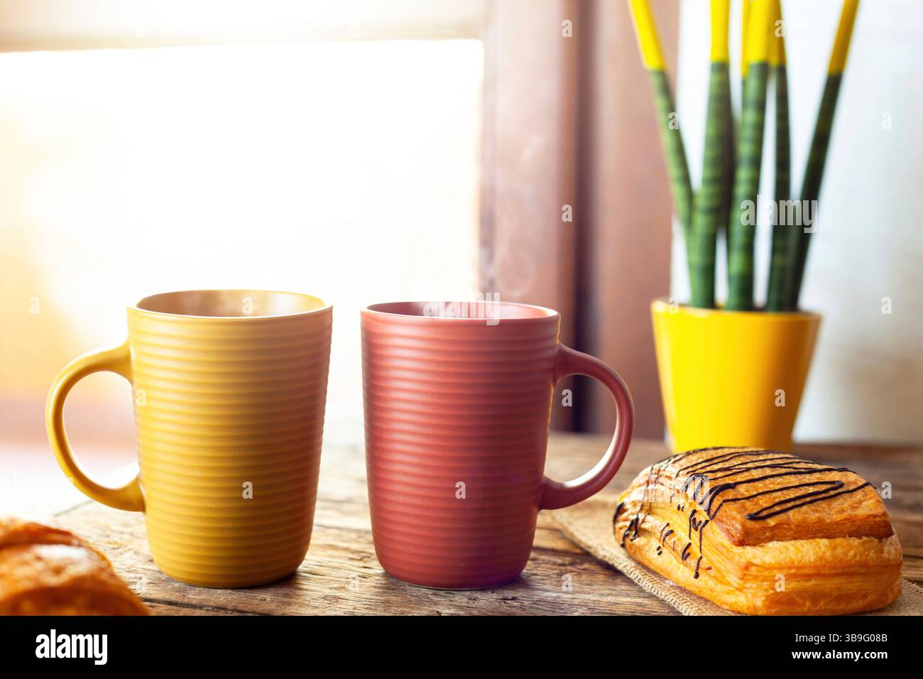 Due tazze fumanti si stendono su un tavolo di legno vicino a una finestra, accompagnate da deliziosi pasticcini, una pianta in vaso gialla e il caldo bagliore della luce del sole mattutino, creando un'atmosfera accogliente per la colazione Foto Stock
