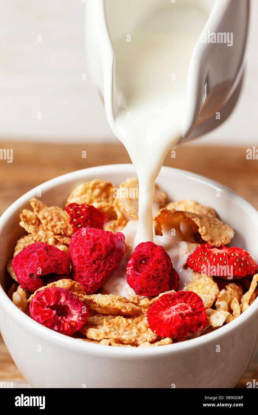 Latte versato da una caraffa bianca in una ciotola di cereali con lamponi e fragole disidratate, per una colazione sana e nutriente Foto Stock