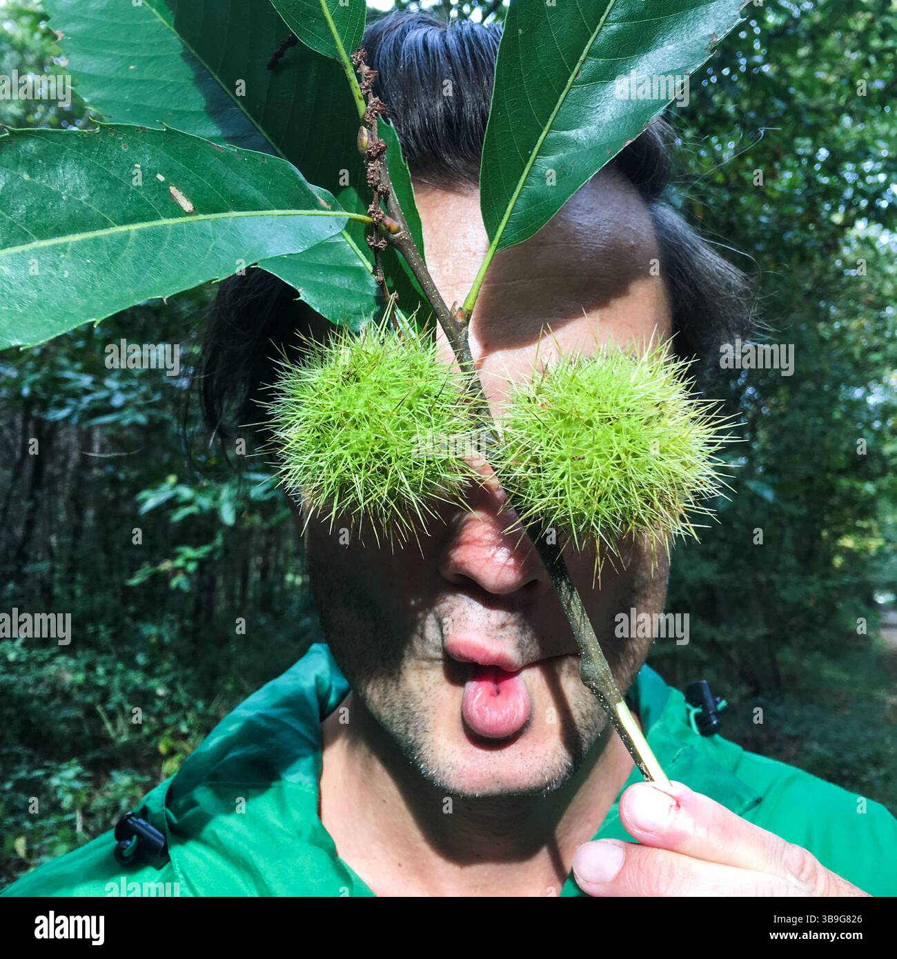 Giovanotto che copre gli occhi con due casse esterne di castagne verdi chiuse, mentre fa una divertente espressione facciale. Divertimento nella foresta autunnale Foto Stock