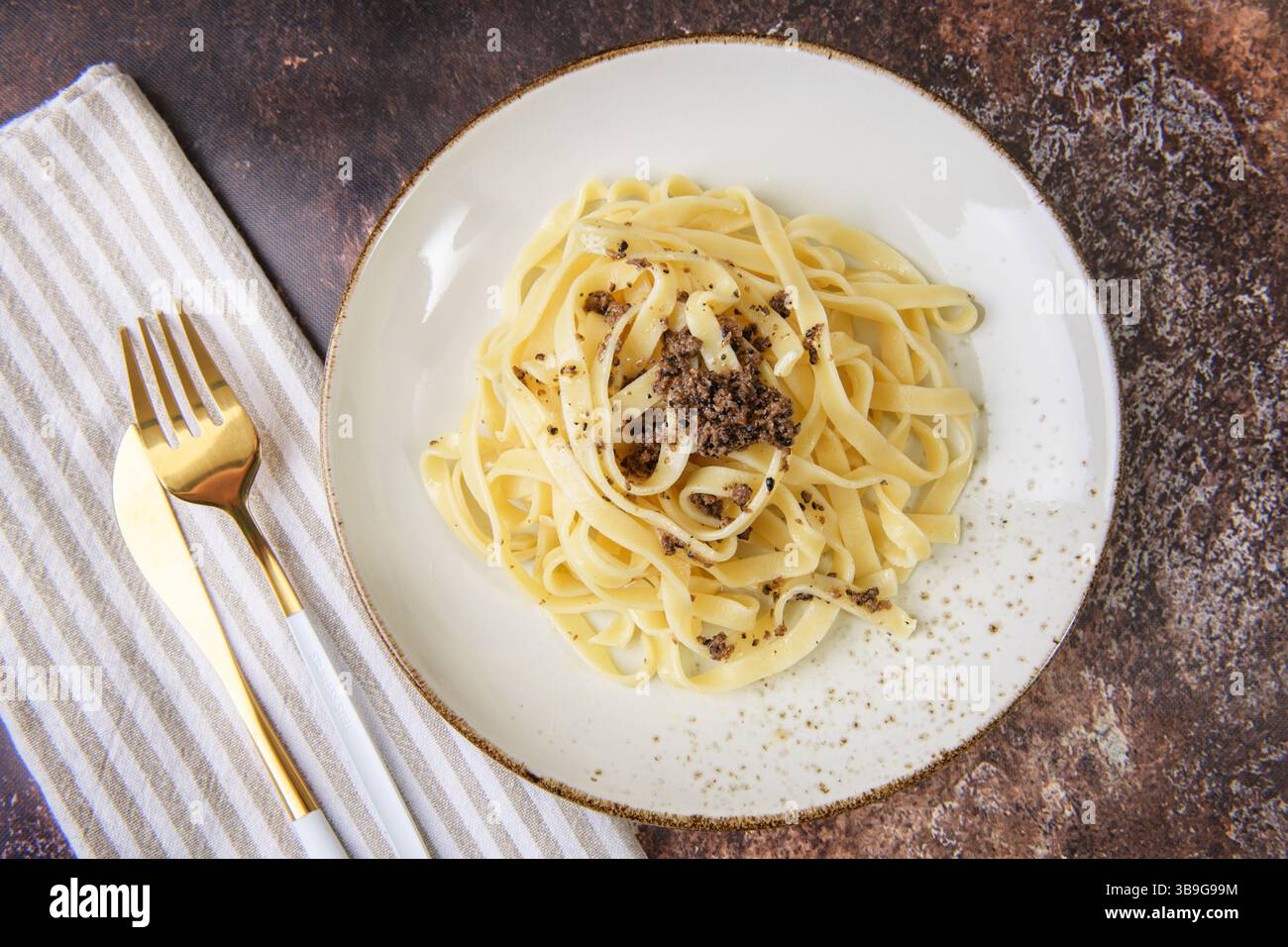 Un piatto di tagliatelle guarnite con tartufo nero, elegantemente collocato su un tavolo rustico. Spara con utensili dorati e un tovagliolo a strisce per dini Foto Stock