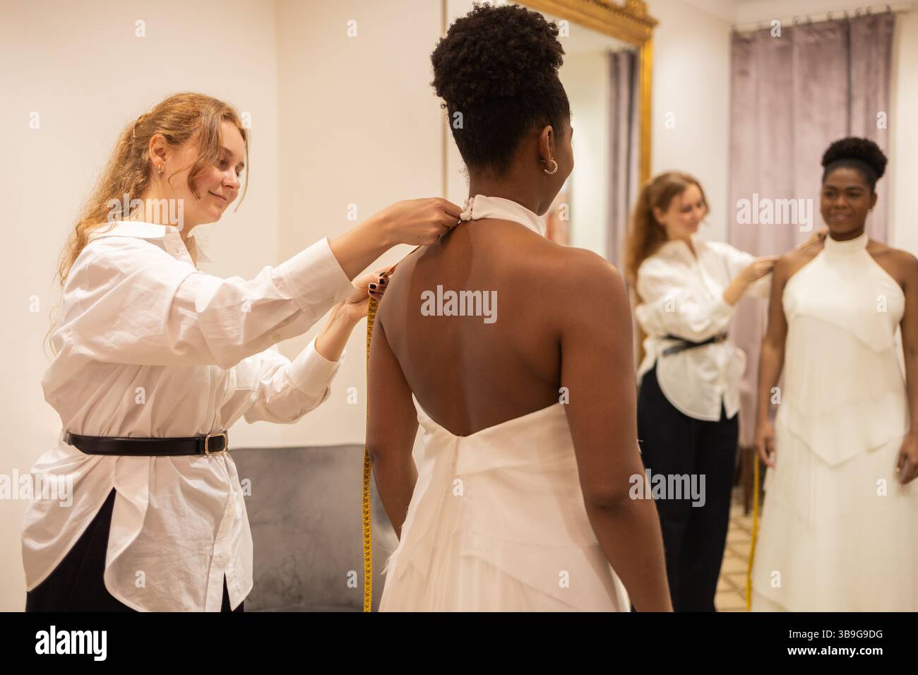 Una sarta adatta un abito da sposa per una sposa in un camerino. La sposa si erge davanti a uno specchio, ammirando l'elegante abito come tocco finale Foto Stock