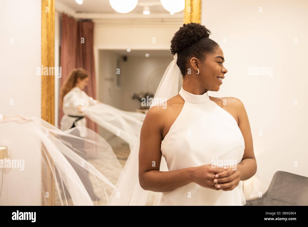 Una sposa sorride elegantemente mentre prova un sofisticato abito da sposa in un negozio nuziale illuminato con calore. Un assistente adatta il suo velo, assicurandosi che ogni deta Foto Stock