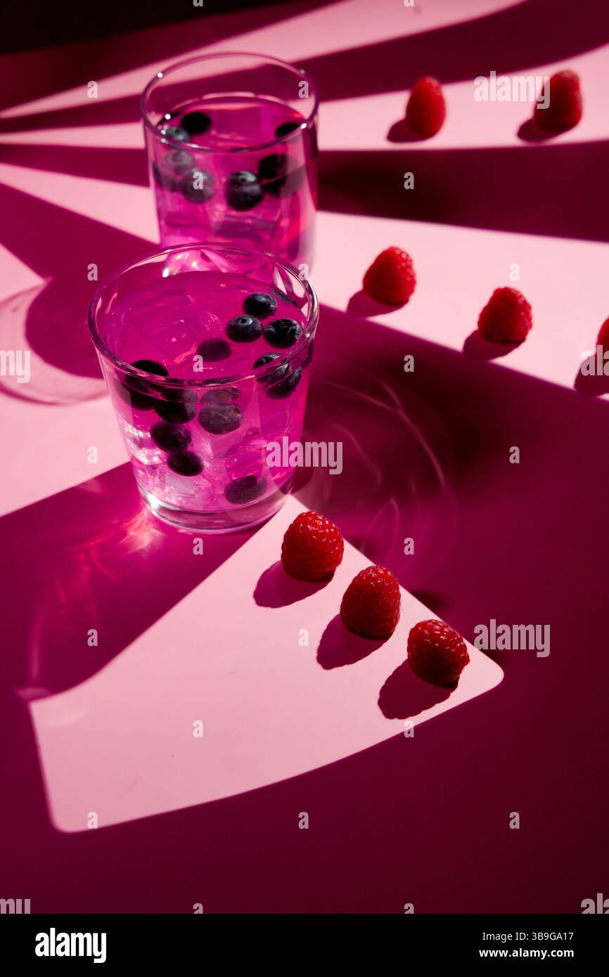 Bevanda rosa vivace con mirtilli in due bicchieri su una superficie rosa. I lamponi sono disposti artisticamente e creano ombre giocose. Perfetto per un re Foto Stock