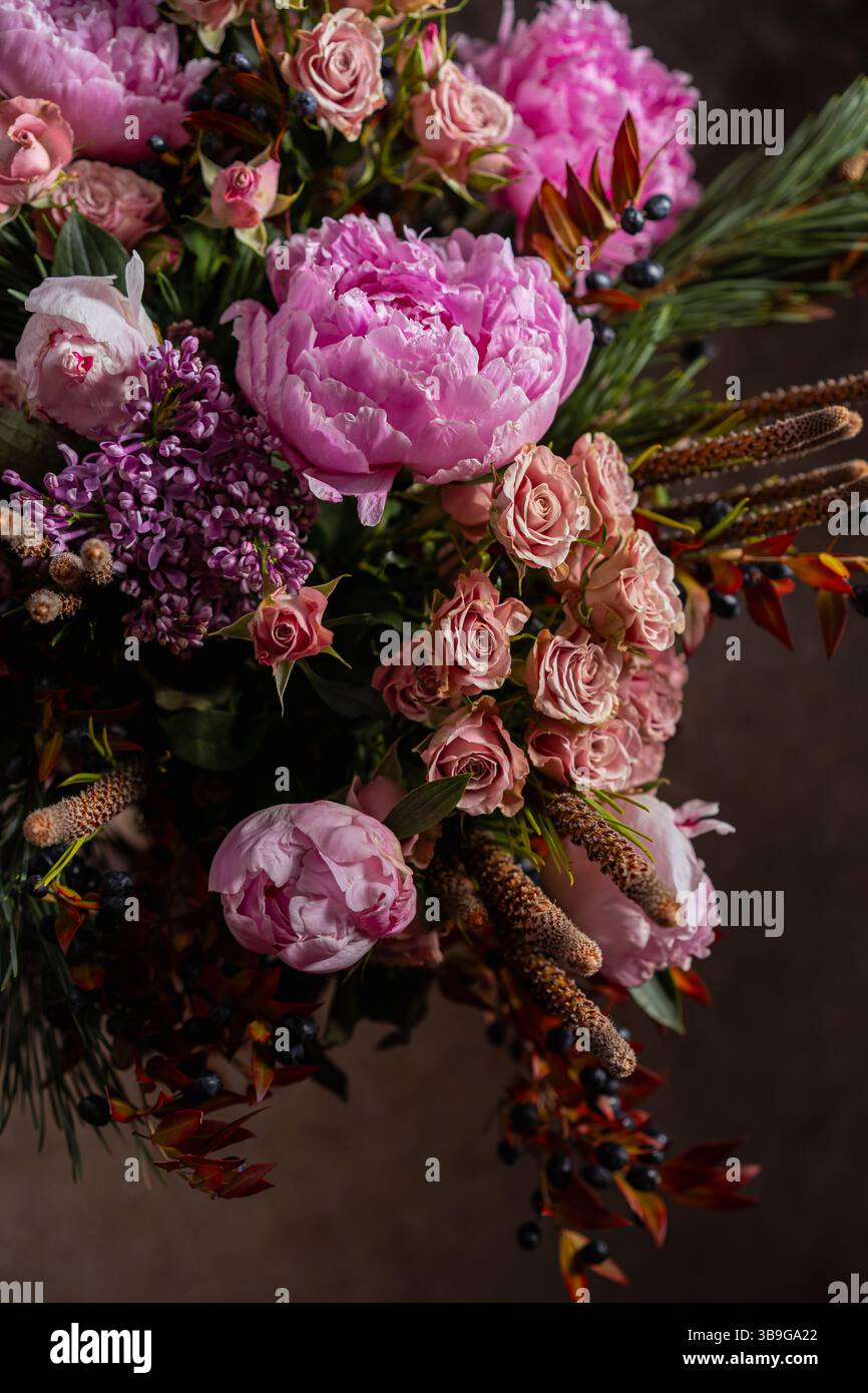 Una lussuosa disposizione floreale caratterizzata da morbide peonie rosa, delicati fiori di lilla e rose rosa, accentuate da bacche scure e fogliame di pino, si mostra Foto Stock