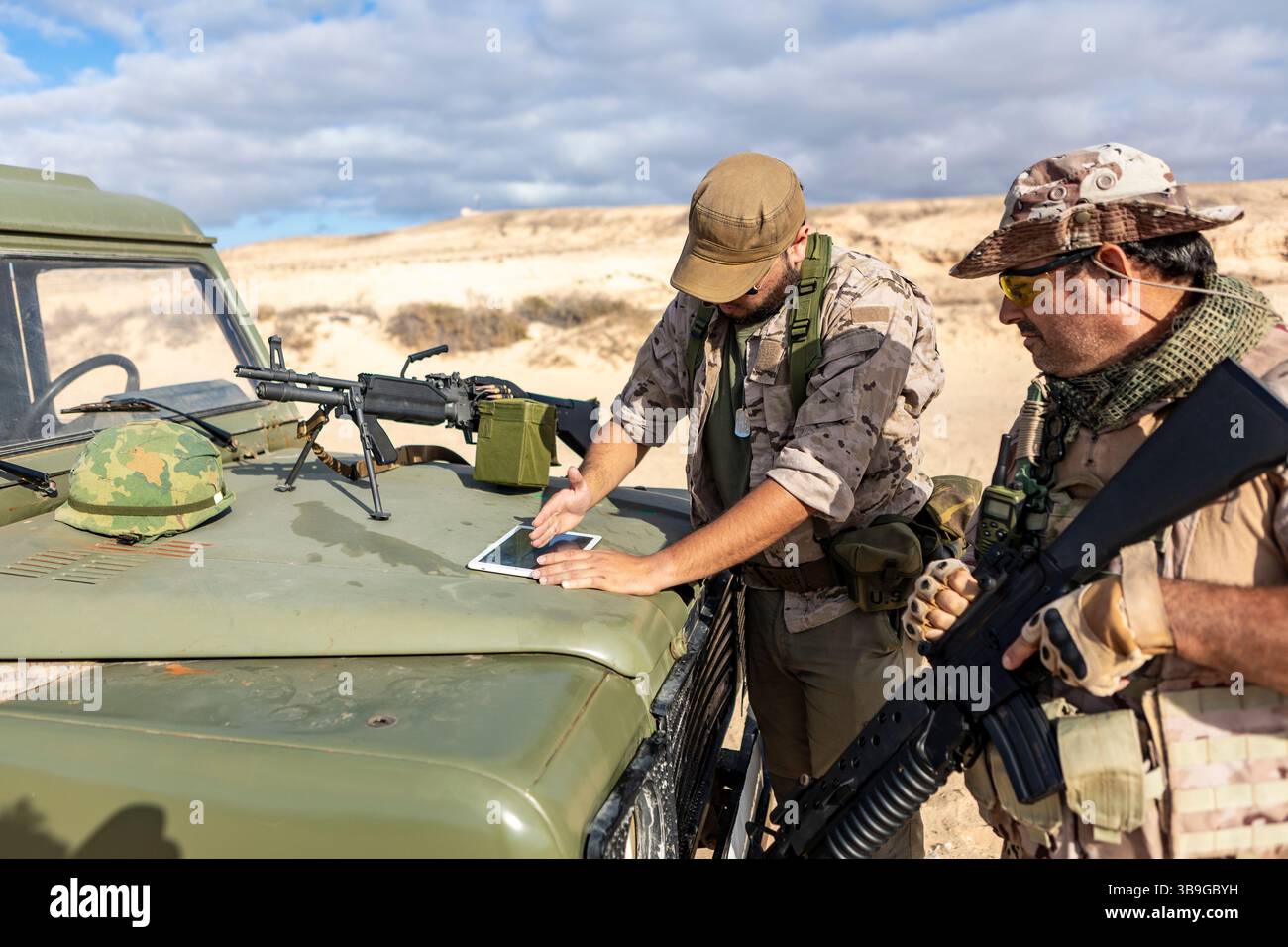 Due soldati militari in equipaggiamento tattico analizzano le mappe digitali su un tablet in cima a un veicolo, preparando la strategia in un ambiente desertico. Attrezzature, inclusi Foto Stock